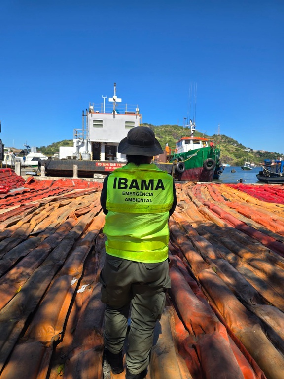 Servidor ou servidora do Ibama, usando colete verde de emergência ambiental, com chapéu, em área portuária, avaliando equipamentos de resposta a emergências com óleo no mar; ao fundo da imagem, aparecem diversas embarcações.  