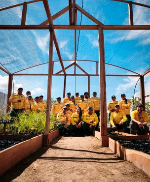 Grupo de brigadistas, mulheres e homens, trajando uniforme amarelo e verde, posa em viveiro com estrutura de madeira e cobertura de tela, cercado por mudas de plantas, sob céu azul.