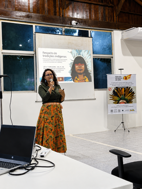 mulher segura microfone e fala sobre respeito às tradições indígenas, conforme informação projetada em tela detrás dela.