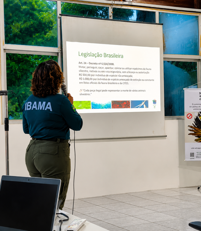 Servidora do Ibama, uniformizada, está de costas para o observador, segurando um microfone e apresentando sobre legislação brasileira. Enquanto fala ao microfone, ela observa uma projeção sobre o assunto em uma tela. 