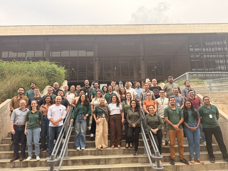 um grupo de aproximadamente 40 servidores posa para uma foto em uma escadaria de cimento, com corrimões centrais, em frente à entrada de um edifício, o qual possui uma estrutura similar a uma grade preta, persiana, como fachada da edificação. Há homens e mulheres no grupo.