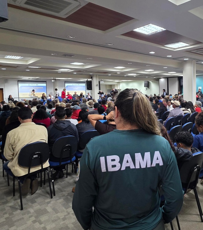 A foto mostra um auditório com mais de 100 pessoas sentadas em cadeiras, voltadas para um grupo de palestrantes, com imagens projetadas atrás deles. Em primeiro plano, há uma servidora do Ibama, de costas, observando a plateia. 
