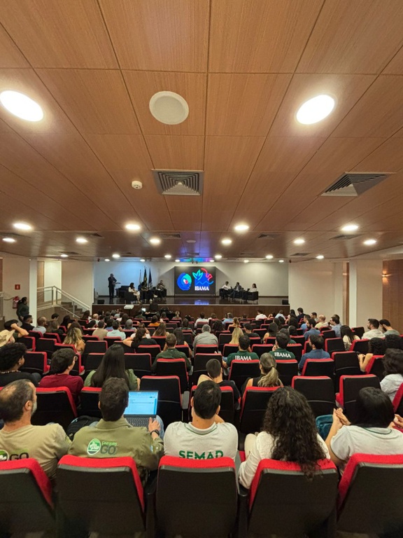 A foto mostra um amplo auditório, cuja plateia conta com cerca de 100 pessoas, sentadas em poltronas de estofado vermelho e fundo preto; o palco do local conta com palestrantes sentados em poltronas quase de frente uma para as outras; a logomarca do Ibama aparece ao centro deles, projetada em um painel.