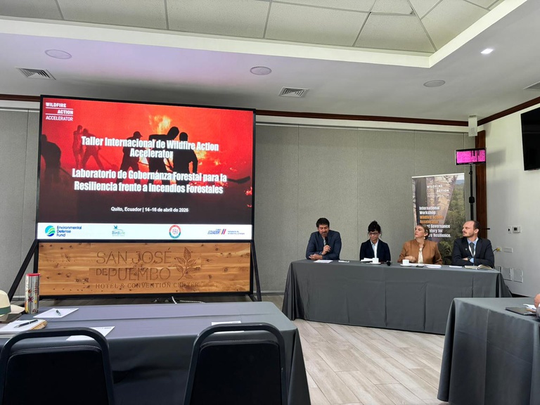 A foto mostra um auditório, com uma mesa posicionada à direita com quatro palestrantes sentados de frente para a plateia, sendo duas mulheres ao centro e um homem em cada ponta da mesa; à esquerda, um painel exibe uma imagem projetada com texto em inglês e espanhol relacionado ao título do evento, bem como com texto menor, abaixo, indicando local e período do evento; atrás dos palestrantes há um banner vertical com imagem de floresta e letras ilegíveis, mas aparentemente relacionadas ao tema; o palestrante homem à esquerda segura um microfone e fala aos presentes; o restante da imagem mostra mesas semelhantes a dos palestrantes, posicionadas em frente ao painel. Não aparecem outras pessoas na imagem.
