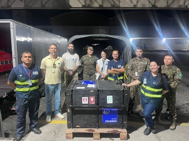  Um grupo de profissionais formado por 6 homens e 3 mulheres envolvidos com o transporte do filhote de onça-pintada está ao lado da gaiola de transporte feita de paredes pretas, na qual está o felino resgatado. Atrás deles, há dois caminhões, estacionados no terminal do aeroporto de Manaus, no Amazonas. A equipe se prepara para colocar a gaiola de transporte dentro do avião que levará a onça até Brasília, no Distrito Federal.  
