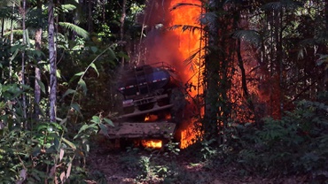 Um caminhão esteira carrega um caminhão tanque de cor preta. Ambos estão em chamas, parados, em uma pequena clareira em meio à floresta densa e verde com características do bioma amazônico.