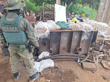 Agente do Ibama, vestido com uniforme e colete de proteção verde, observa equipamentos e materiais apreendidos em uma área de garimpo ilegal. Sobre uma estrutura metálica estão diversos tubos e peças utilizados na atividade de mineração. O ambiente é aberto, com ferramentas e sucata espalhadas ao fundo. 