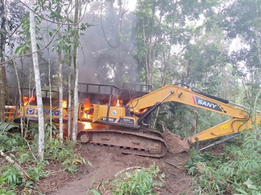 Escavadeira amarela em chamas dentro de uma área de floresta amazônica. Fumaça escura sobe entre as árvores enquanto o equipamento é destruído durante ação de fiscalização ambiental em região de mata fechada. 