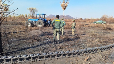 agentes ambientais federais do Ibama estão em uma área devastada pelo fogo, abordando pessoas que utilizam um trator azul no momento da fiscalização; ao fundo, há uma vegetação seca e, próximo dela, uma viatura do Ibama; mais atrás, há um relevo de serra; em primeiro plano, há um “correntão” no chão, provavelmente usado para desmatar áreas.