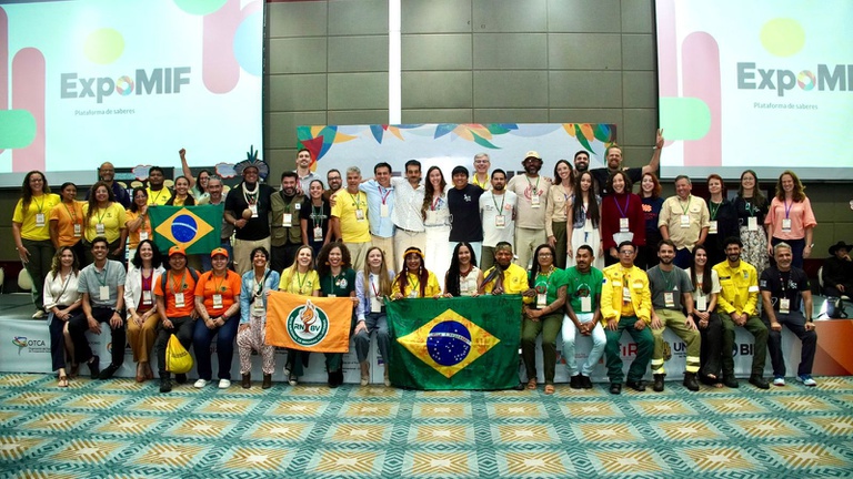 Foto colorida, em ambiente interno de auditório. Um grupo numeroso de participantes está posicionado em várias fileiras para uma foto coletiva. As pessoas vestem roupas variadas, incluindo camisetas institucionais, camisas, coletes e uniformes operacionais. Na parte frontal do grupo, há bandeiras estendidas, entre elas a bandeira do Brasil. Ao fundo, um painel com a identidade visual da ExpoMIF aparece projetado em duas telas laterais, com cores vivas e o nome do evento. O piso tem padrão geométrico e o espaço é amplo e bem iluminado.
