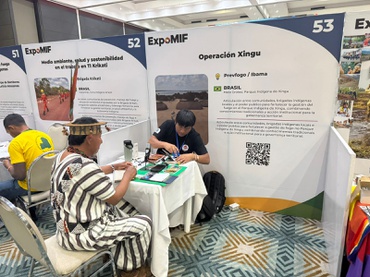Foto colorida em ambiente interno de feira ou congresso. Dois participantes estão sentados frente a frente em uma mesa branca, conversando e manuseando materiais impressos. Ao redor da mesa, há painéis expositivos da ExpoMIF com textos e imagens sobre boas práticas de Manejo Integrado do Fogo. Um dos painéis destaca a “Operação Xingu”, do Prevfogo/Ibama, com menção ao Brasil e um código QR visível. O espaço é bem iluminado, com piso de padrão geométrico em tons de azul e cinza, e outros estandes aparecem parcialmente ao fundo
