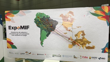 Foto colorida de um painel expositivo em ambiente interno. O painel mostra um mapa estilizado da América do Sul e da Europa, com marcações feitas por alfinetes coloridos interligados por linhas de diferentes cores. As linhas cruzam o mapa, conectando pontos entre países amazônicos e outras regiões, representando acordos e parcerias estabelecidos durante a ExpoMIF. À esquerda, aparece o logotipo do evento e o texto “ExpoMIF — Plataforma de saberes y soluciones por una amazonia más resiliente al fuego”. Na parte inferior do painel, há logotipos de instituições parceiras e apoiadoras