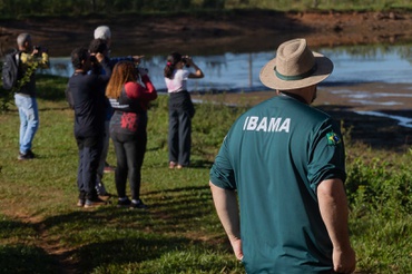 A foto mostra um servidor do Ibama de costas, uniformizado e de chapéu de palha, e mais adiante dele, um grupo de pessoas. Todos estão com a atenção voltada para a paisagem à frente, alguns segurando binóculos e outros apontando para algum objeto de interesse. O local em todos estão, mostrado na imagem, contém uma grama baixa e uma área alagada, similar às margens rasas de um rio. 