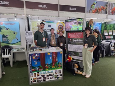 A imagem mostra servidores do Ibama posando para uma foto em um estande do Instituto dentro do evento Avistar Brasil 2026; há um banner ao lado de uma servidora sobre o Programa Caminhos da Biodiversidade; sobre a bancada do stande, há um material de educação ambiental e, colado à sua frente, um banner sobre fauna com fotos de animais silvestres diversos.