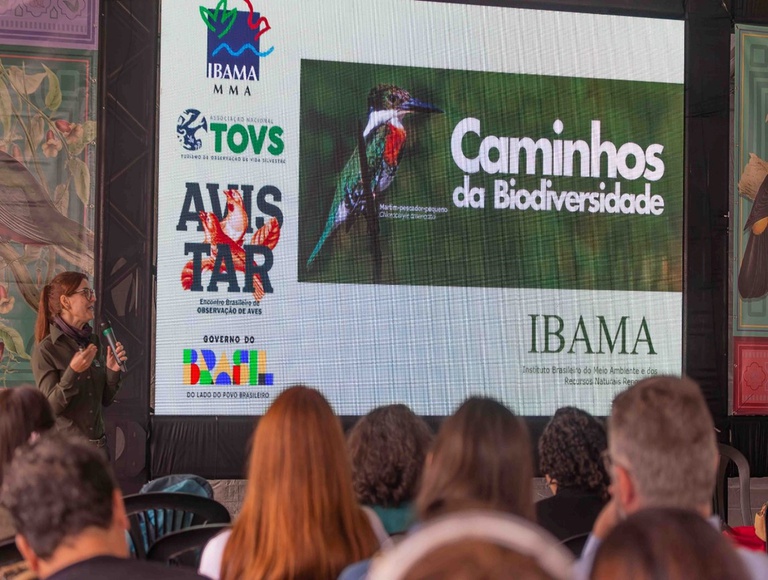 A imagem mostra um grande telão com imagem contendo apresentando sobre o Programa Caminhos da Biodiversidade, do Ibama, e logomarcas de instituições envolvidas e do governo federal