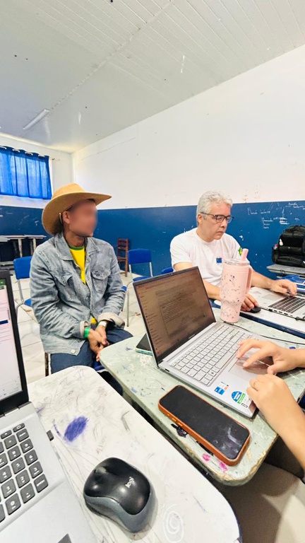 A foto mostra um homem de jaqueta e chapéu sentado em frente a uma mesa com um notebook, o qual é manuseado por alguém. Ao lado do homem, há um servidor do Ibama, também sentado e trabalhando em um notebook; eles estão em uma sala de uma escola.