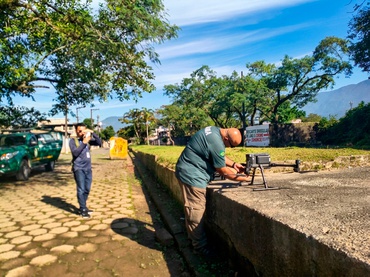 a imagem mostra uma rua residencial pavimentada com pedras poligonais, com uma calçada larga e bem elevada do lado direito; há uma viatura do Ibama estacionada na calçada esquerda da rua; um servidor do Ibama, uniformizado, maneja um equipamento similar a um drone, que está parado na calçada elevada; outro servidor mais ao fundo da imagem, próximo à viatura, registra a ação, com celular, do servidor que maneja o equipamento. 
