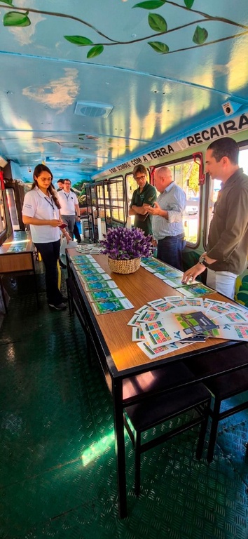 A foto mostra o interior de um ônibus, que não possui cadeiras; o piso é de aço de cor verde, e o teto é branco brilhante; há uma mesa com tampo de madeira e armação de ferro preto, que tem sobre ela materiais educativos semelhantes a folhetos e um jarro de plantas no centro; no local estão servidores de um lado e do outro da mesa e também ao fundo, além de outra mesa menor e uma prateleira.