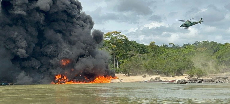 Foto de operacão do Ibama em uma praia, com uma faixa de água, uma faixa de areia e uma mata, na faixa de areia à esquerda há equipamentos sendo inutilizados com fogo e uma nuvem preta do material queimado que sobe ao céu, no lado inferior direito há um pequeno barco com motor atracado na praia, e acima à direita na faixa de céu após a mata avista-se o helicóptero do Ibama vigiando a ação.