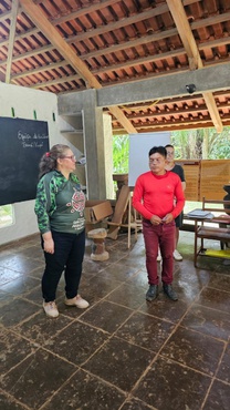 Sala com piso de cerâmica, estrutura de madeira e abertura para área externa com vegetação. Em primeiro plano, uma mulher e um participante indígena estão em pé, voltados um para o outro, durante atividade formativa. Ao fundo, há outra participante em pé e, na parede, um quadro negro com anotações e cartazes informativos afixados.