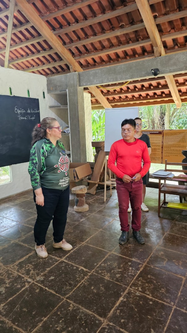Sala com piso de cerâmica, estrutura de madeira e abertura para área externa com vegetação. Em primeiro plano, uma mulher e um participante indígena estão em pé, voltados um para o outro, durante atividade formativa. Ao fundo, há outra participante em pé e, na parede, um quadro negro com anotações e cartazes informativos afixados. 