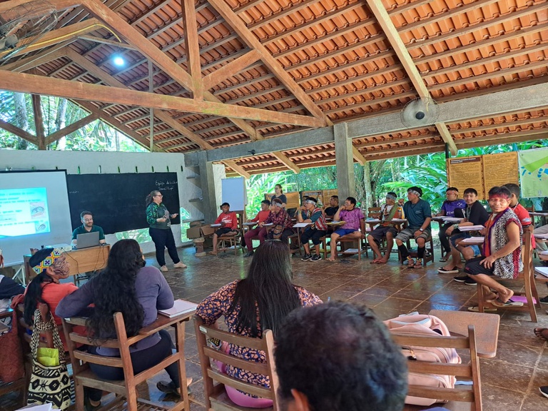 Um ambiente interno de uma sala ampla, com estrutura de madeira e telhado aparente, aberta para a área externa com vegetação ao redor. Cerca de vinte pessoas, entre indígenas e facilitadores, estão sentadas em cadeiras dispostas em semicírculo. À frente, uma mulher branca com cabelos parcialmente grisalhos está em pé conduzindo a atividade, enquanto um homem opera um computador conectado a um projetor, que exibe imagens em uma tela grande. Os participantes observam atentamente, alguns com cadernos nas mãos. Ao fundo, há um quadro negro e painéis informativos fixados na parede. 