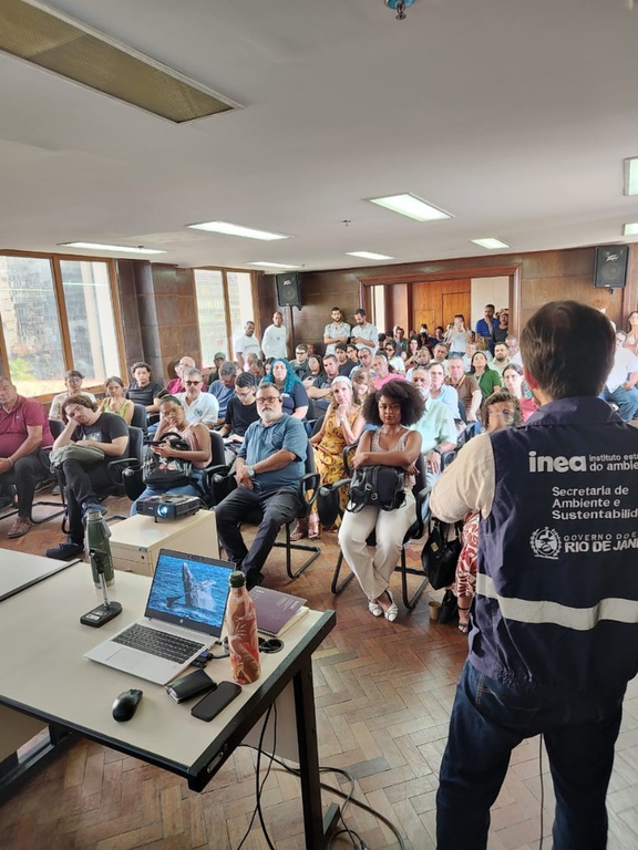 A foto mostra uma plateia com cerca de 60 pessoas sentadas em cadeiras em um pequeno auditório. O local tem janelas grandes de vidro do lado esquerdo, e o piso é de taco. Há um palestrante falando ao público com colete do Inea-RJ e, ao lado dele, uma mesa com um notebook e outros itens.