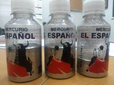 Em primeiro plano, três frascos plásticos transparentes, com tampa branca de rosca, estão alinhados sobre uma mesa de madeira clara. Os recipientes contêm mercúrio metálico, de aspecto prateado e brilhante, ocupando aproximadamente a metade de cada frasco. Nos rótulos, predominam as cores preta, branca e vermelha, com a inscrição
