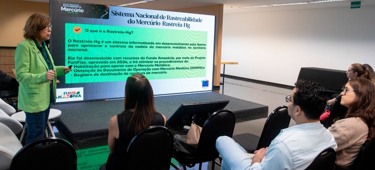 Em uma sala de eventos, uma mulher em pé, à esquerda da imagem, faz uma apresentação para um grupo de pessoas sentadas em cadeiras voltadas para um telão. Ela veste blazer verde-claro, blusa escura, calça jeans e óculos, e segura um controle remoto enquanto gesticula durante a exposição. No telão, aparece o título "Sistema Nacional de Rastreabilidade do Mercúrio – Rastreia-Hg", acompanhado de um texto explicativo sobre o sistema informatizado desenvolvido pelo Ibama para aprimorar o controle da cadeia do mercúrio metálico no Brasil. Também são exibidos os logotipos do Fundo Amazônia e do Ibama. Em primeiro plano, participantes assistem atentamente à apresentação. O ambiente é amplo, com piso escuro, paredes claras, iluminação por luminárias no teto e uma pequena plataforma onde está instalado o telão.