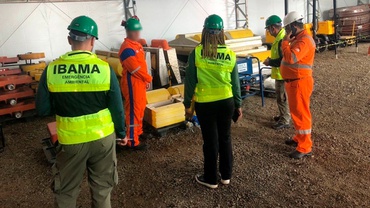A foto mostra uma espécie de depósito de materiais ou equipamentos não identificados, com chão arenoso; no local estão três servidores do Ibama (dois homens e uma mulher), de costas, utilizando coletes verdes fluorescentes com o nome “IBAMA” e, abaixo, escrito “Emergência Ambiental” em letras menores; eles conversam com dois homens com uniformes laranjas; todos as pessoas utilizam capacetes de proteção.