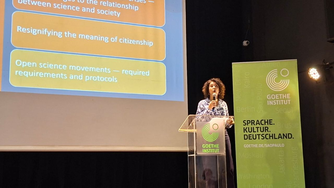 Sarita Albagli, pesquisadora do Ibict, fala ao microfone atrás de um púlpito transparente com o logotipo do Goethe-Institut. Ao fundo, uma tela projeta slides em inglês sobre ciência aberta e cidadania. À direita, um painel verde exibe a identidade visual do Goethe-Institut.