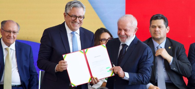 Foto do presidente Lula com o Ministro Alexandre padilha, ambos estão sorrindo segurando uma pasta com documentos
