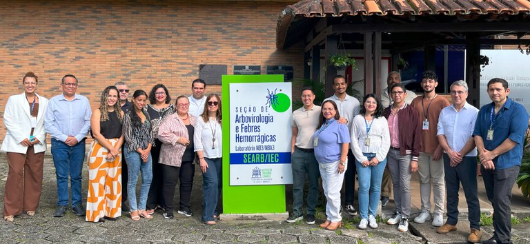 Grupo diverso de pessoas posa sorrindo em frente à placa da Seção de Arbovirologia do IEC.