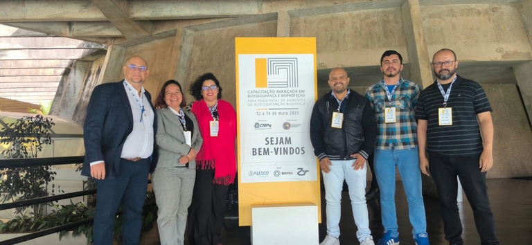 Seis pessoas sorrindo ao lado de placa de evento sobre biossegurança, em ambiente moderno de concreto.