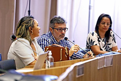 Três pessoas sentadas em mesa de evento. Homem de óculos fala ao microfone; mulheres escutam atentas.