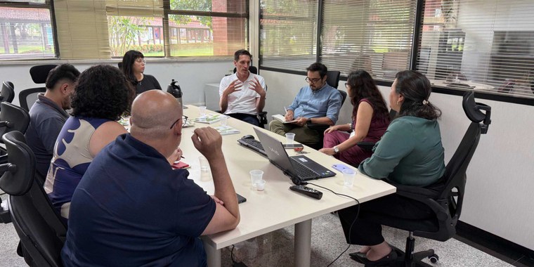 Reunião em sala; oito pessoas sentadas em mesa, homem ao centro gesticula.