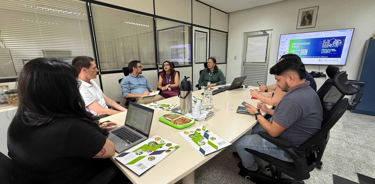 Visão lateral da mesma reunião; pasta IEC e café sobre a mesa.