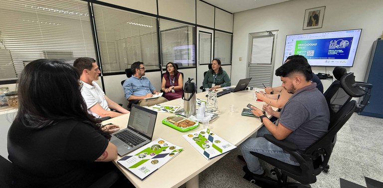 Visão lateral da mesma reunião; pasta IEC e café sobre a mesa.