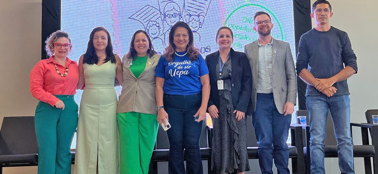 Sete pessoas sorrindo lado a lado em palco, diante de telão colorido com ilustração e textos.