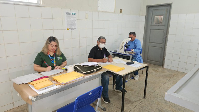 Três profissionais trabalham em laboratório de campo; um usa microscópio e dois preenchem papéis.