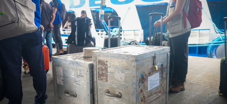 Pessoas embarcando com baús metálicos em frente a uma embarcação branca.