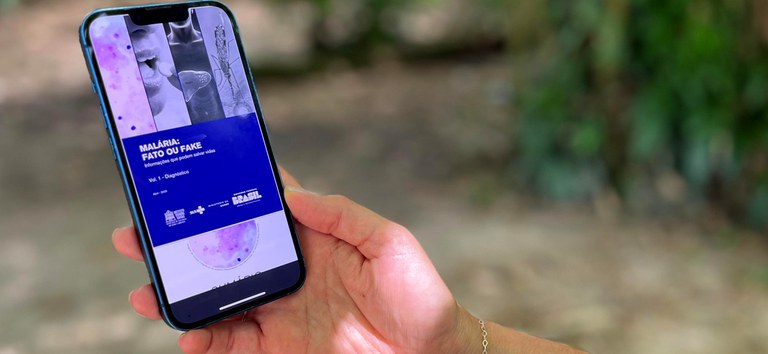 Pessoa segura celular com e-book “Malária: Fato ou Fake” aberto na tela, em ambiente externo.