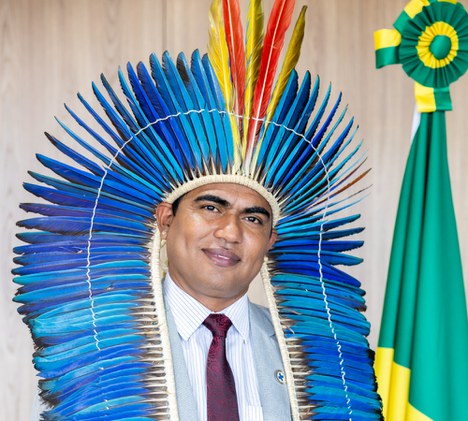 Homem indigena sorrindo para foto, ele possui uma indumentária indigena e ao fundo a bandeira nacionaç