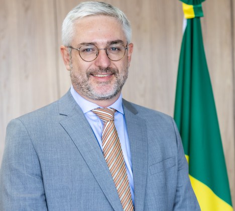 Foto de homem branco sorrindo, com a bandeira do Brasil ao fundo