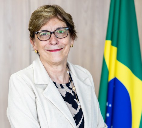 Foto de senhora branca sorrindo e ao fundo a bandeira do Brasil