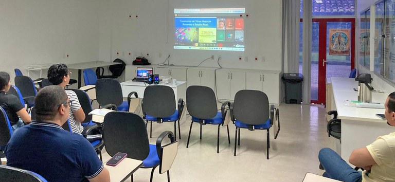 Sala com cadeiras azuis, poucas pessoas assistem a palestra projetada na parede branca.