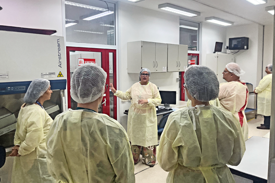 Um grupo de pessoas em um laboratório, vestindo jalecos e toucas, ouvindo uma mulher explicando algo.