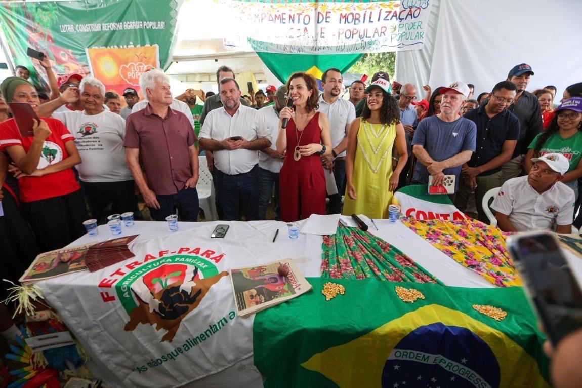 Pessoas em pé ao redor de uma mesa e ministra ao centro com microfone.