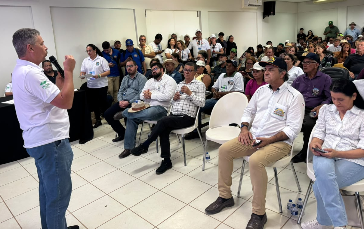 Um homem fala ao microfone, em pé, para um grupo de pessoas sentadas em uma ampla sala.