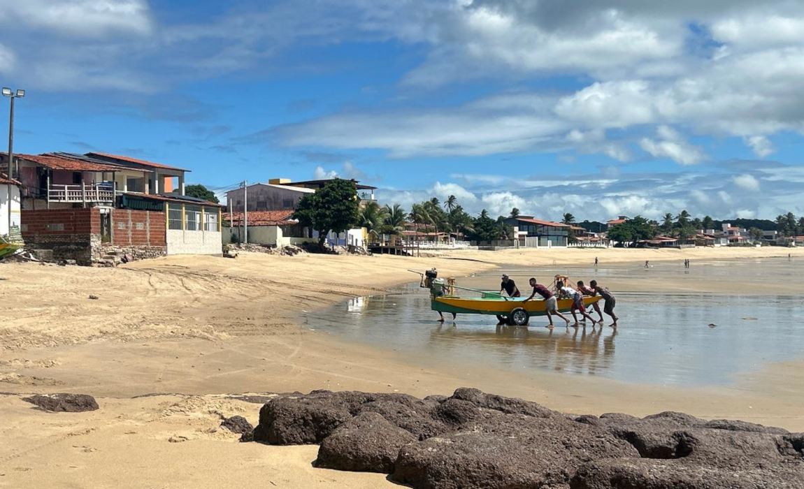 Pescadores empurram um barco saindo do mar e chegando a uma aldeia com várias casas.
